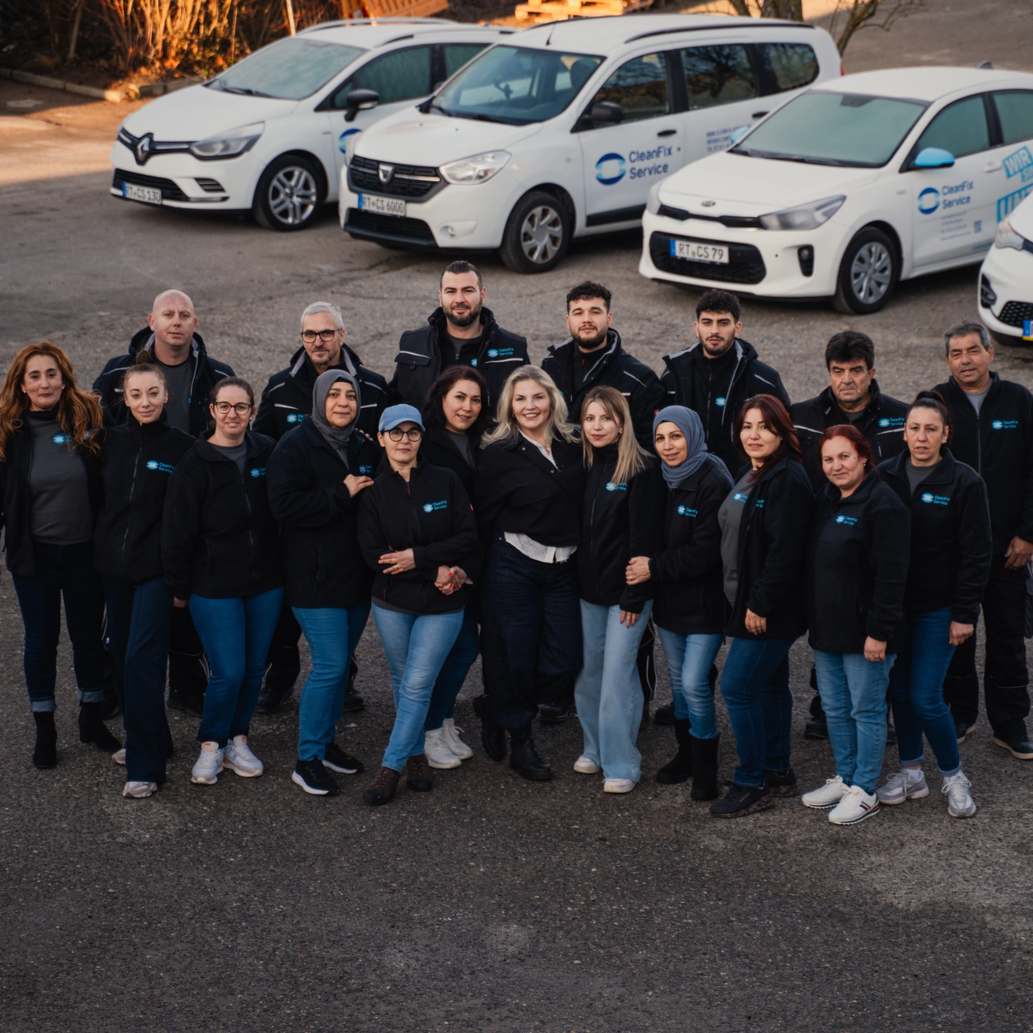 Team Das CleanFix Service Team in dunklen Arbeitsuniformjacken mit Logo, aufgestellt vor vier weißen Firmenfahrzeugen mit CleanFix Service Beschriftung.